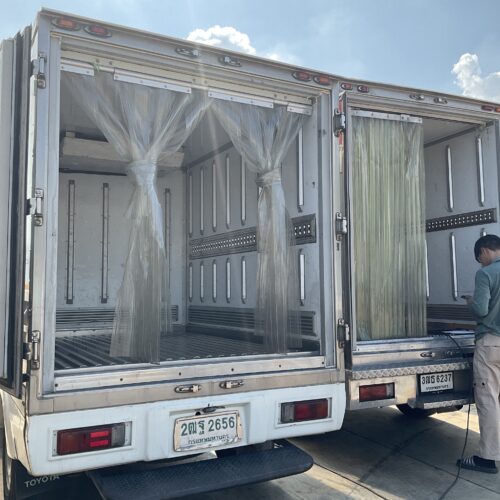 Open rear of a metal cargo truck with clear plastic curtains and vertical support rails, parked under a bright sky.