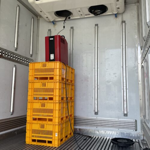 Interior of a refrigerated trailer with stacked yellow plastic crates and a red electrical unit mounted on a rack, cooling unit overhead