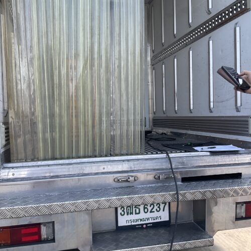 Open rear of a metal cargo truck with corrugated walls; a hand holds a smartphone on the right, papers on the diamond-plate step, Thai license plate visible.