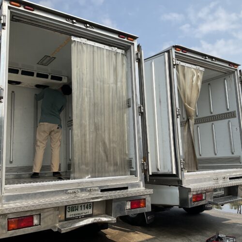 Two open-back cargo trucks with a worker unloading inside one truck bay; metallic doors and reflective surfaces visible against a blue sky.
